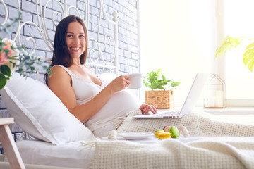 Smiling pregnant woman breakfasting in bed with laptop