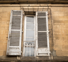 Old windows with weathered shutters in a facade of a kind of mansion with a forged ornamental fence, viewing point is from below. Coloring is somewhat bleak and gray. Style is old French.