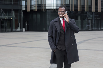 Horizontal photo of handsome dark-skinned African businessman pictured outdoors in street on his way from or to business meeting, discussing important issues with clients or colleagues on phone - Powered by Adobe