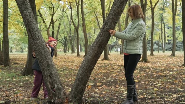 Little cute girl and her mother playing hide and seek in the autumn park, hiding behind the trunks of trees. Slow motion 