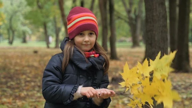 Cute little girl is covering her face and waving a maple tree branch with yellow leaves in autumn park. Slow motion.