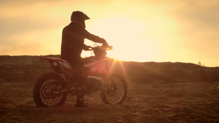 Plakat Professional FMX Motorcycle Rider Rests on His Bike and Overlooks Hard Sandy Off-Road Terrain. Sun is Setting.