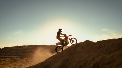 Side View Footage of the Professional Motocross Motorcycle Rider Driving on the Dune and Further Down the Off-Road Track. It's Sunset and Track is Covered with Smoke/ Mist.
