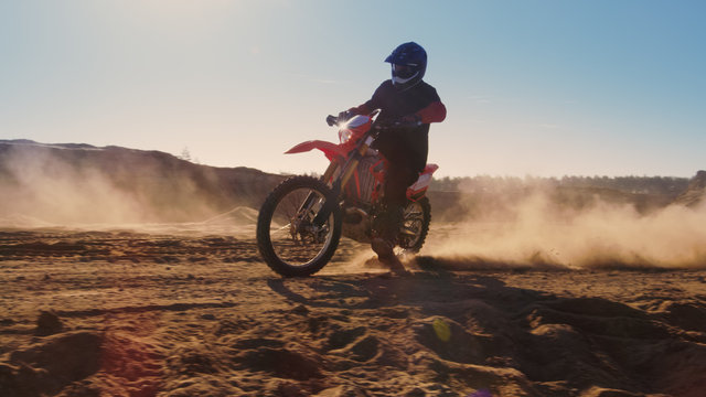 Side View Footage of the Professional Motocross Motorcycle Rider Driving on the Dune and Further Down the Off-Road Track. It's Sunset and Track is Covered with Smoke/ Dust/ Dirt.