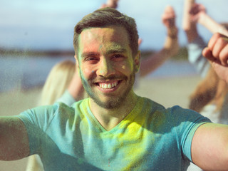 Selfie Portrait of a Muscular Man Celebrating of Holi Festival With His Friends. His Face and Clothes are Covered with Colorful Powder. People Dancing in the Background.