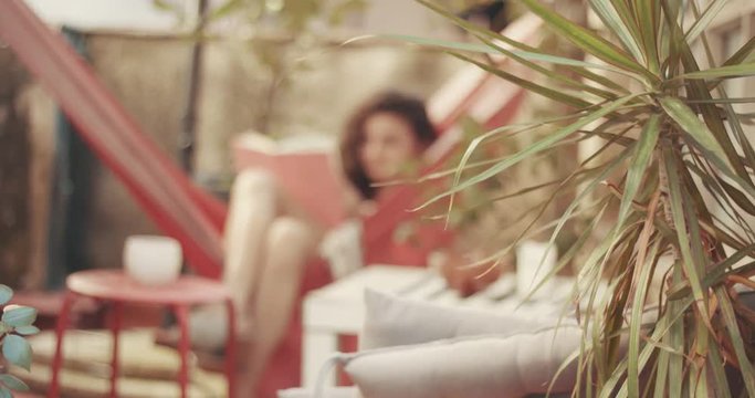 Pretty red-haired young woman in floral dress reads a book in a hammock in a small urban patio garden