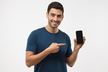 Shot of hansome guy in blue t-shirt isolated on grey background, presenting smart phone and pointing with finger at blank black screen with copyspace for ads