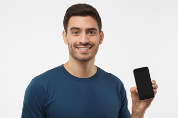 Closeup of young handsome man smiling isolated on grey background, holding and showing blank smartphone with copyspace for advertisement of applications, websites, goods and services