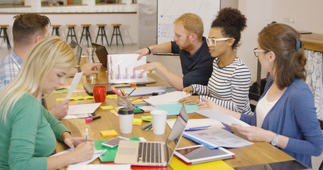 Diverse group of young people gathering at table in modern office and working with documents and gadgets creating new plan.