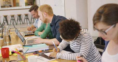 Group of diverse employees sitting together at long table with technology and papers and working...