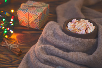 Big cup of hot cocoa with marshmallow, gift box, cinnamon and warm blanket on an old vintage wooden and Christmas light. Cozy christmas or autumn arrangement.