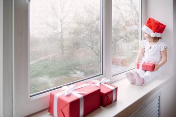 Fototapeta premium little girl in Santa hat sits on window sill and holding gift