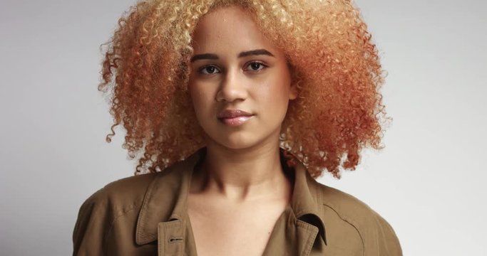 Sensual flirty young mixed race female model with blond red afro hair in a long beige trenchcoat on white background
