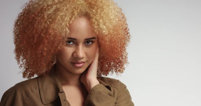 Sensual flirty young mixed race female model with blond red afro hair in a long beige trenchcoat on white background