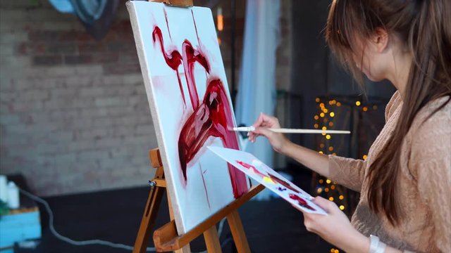 Very talanted artist is drawing three pink flamingos on an easel. She is working in an art studio alone.