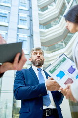Fototapeta premium Low angle view of confident business partners discussing details of mutually beneficial cooperation while standing at spacious office lobby, assistant manager taking necessary notes