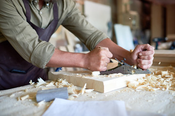Side view closeup of strong male hands shaving piece of wood with plane tool in carpenters workshop making furniture