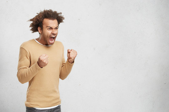 Furious outaged man shouts in anger, keeps fists, going to defent himself in fight with criminals. Desperate dark skinned mixed race male with bushy curly hair shows his power, clenches fists
