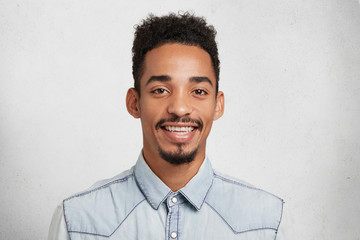 Indoor shot of glad young bearded man and mustache, wears denim shirt, smiles happily, rejoice successful day, being promoted at work, poses against white background. Facial expressions concept