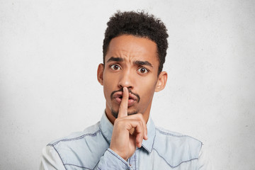 Serious dark skinned male keeps finger on lips, asks to be silent and not interrupt him, isolated...