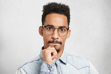 Portrait of mixed race serious focused bearded man with Afro hairstyle, keeps hand on chin, pressses lips, listens attentively someone talk or presentation, isolated over white studio background