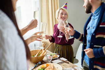 Group of happy young people wearing holiday caps celebrating Birthday with friends clinking champagne glasses during dinner party at home