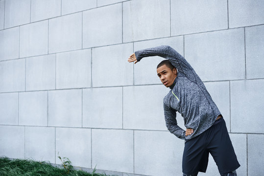 Isolated outdoor portrait of confident young dark-skinned sportsman in stylish outfit bending sideways, doing stretching and warm up exercises, preparing his mucles for workout. Sports and motivation