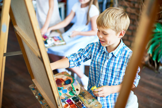 Portrait of happy  little boy painting on easel during art class in sunlit studio enjoying work