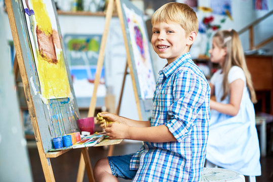 Side view portrait of little boy fingerpainting house on easel in art studio and looking at camera smiling happily