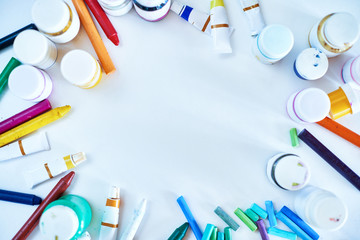 Background image of art supplies: colorful pencils, crayons, pastels and gouache cans scattered around blank white sheet of paper, copy space