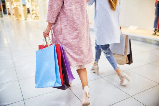 Low section portrait of two young women wearing coats and high heels walking in shopping mall holding paper bags with purchases