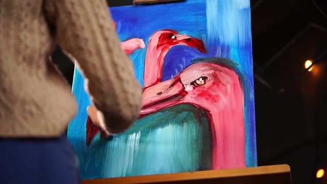 Painter is standing in front of almost finished picture of three caribbean flamingos in contemporary style. She is making final touches on a blue background, back view.