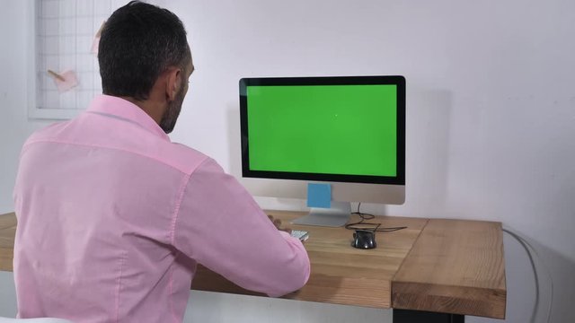 Back view man sitting at the desk with computer green screen. Male with black gray hair typing on keyboard entering data in room with white wall