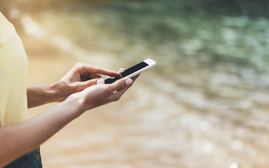 Hipster girl hold on smart phone gadget in sand coastline, mock up blank screen. Traveler using in female hand mobile on background beach seascape horizon. Tourist look on blue sun ocean