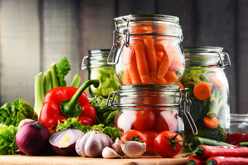 Jars with marinated food and raw vegetables on cutting board