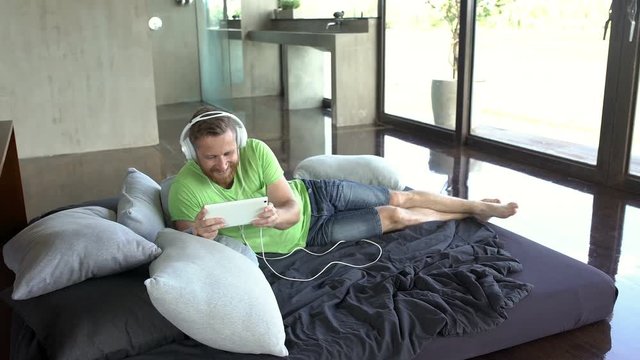 Man listening music and playing game on tablet while lying on bed
