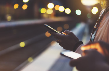 Enjoying travel. woman waiting on station platform on background light moving train using smart phone in night. Tourist texting message and plan route of stop railway, railroad transport, bokeh