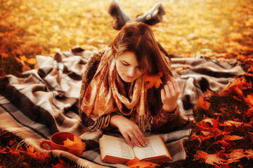 Beautiful young brunette sitting on a fallen autumn leaves in a park, reading a book