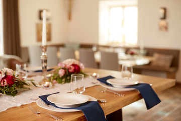 Tischdekoration mit kleinen Blumen, Kerzen und einem weisen Tischläufer auf einem Holztisch in einem großen offenem Raum. Gedeckt sind weise Teller auf blauen Servietten mit silbernen Geschirr.