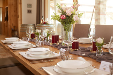 Fototapeta na wymiar Tischdekoration mit Blumenstrauß und roten Kerzen auf einem grauen Tischläufer auf einem Holztisch in einem großen offenem Raum. Gedeckt ist klassisches weises Geschirr.