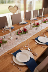 Tischdekoration mit kleinen Blumen, Kerzen und einem weisen Tischläufer auf einem Holztisch in einem großen offenem Raum. Gedeckt sind weise Teller auf blauen Servietten mit silbernen Geschirr.