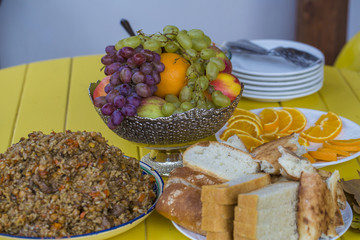 on the yellow table stood a bowl of plov. There are fruits in the vase. Bread is cut into pieces.