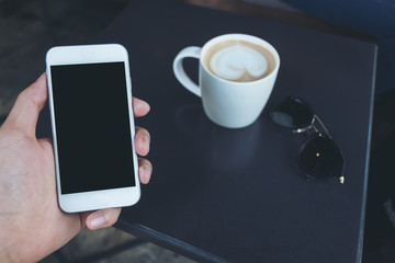 Mockup image of a man's hand holding white mobile phone with blank black screen , coffee cup and sunglasses on a table in cafe background