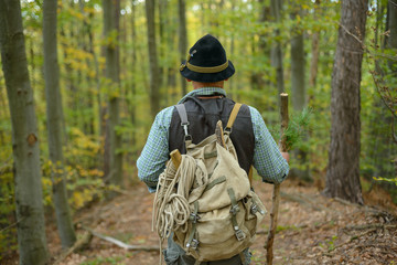 Naklejka premium An old man in a hat with a backpack on holiday in the mountains. The elder is leaning against a stick in the autumn forest. 