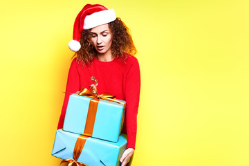 Young Santa woman in christmas hat wearing red sweater with deer smiling holding christmas gifts and having fun laughs happy rejoices touches on yellow background in the studio