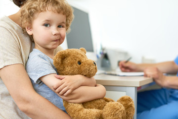 Portrait of cute child sitting on mothers lap in doctors office hugging plush teddy bear  and looking at camera, copy space