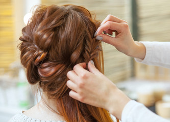 Fototapeta premium Beautiful, red-haired girl with long hair, hairdresser weaves a French braid, in a beauty salon. Professional hair care and creating hairstyles.