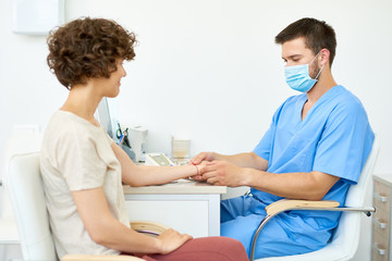 Fototapeta premium Portrait of smiling young woman talking to doctor wearing face mask and holding her arm for blood test