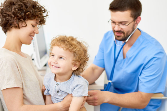 Portrait of happy little boy  sitting on mothers lap in doctors office while pediatrician listening to breathing using stethoscope