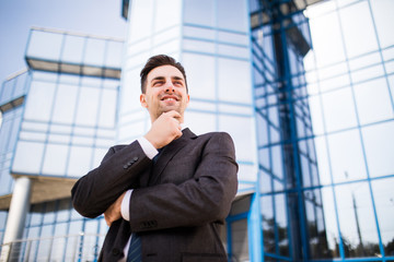 Confident thoughtful businessman in a suit holding hand on chin and looking away while standing outdoors with office building in the background